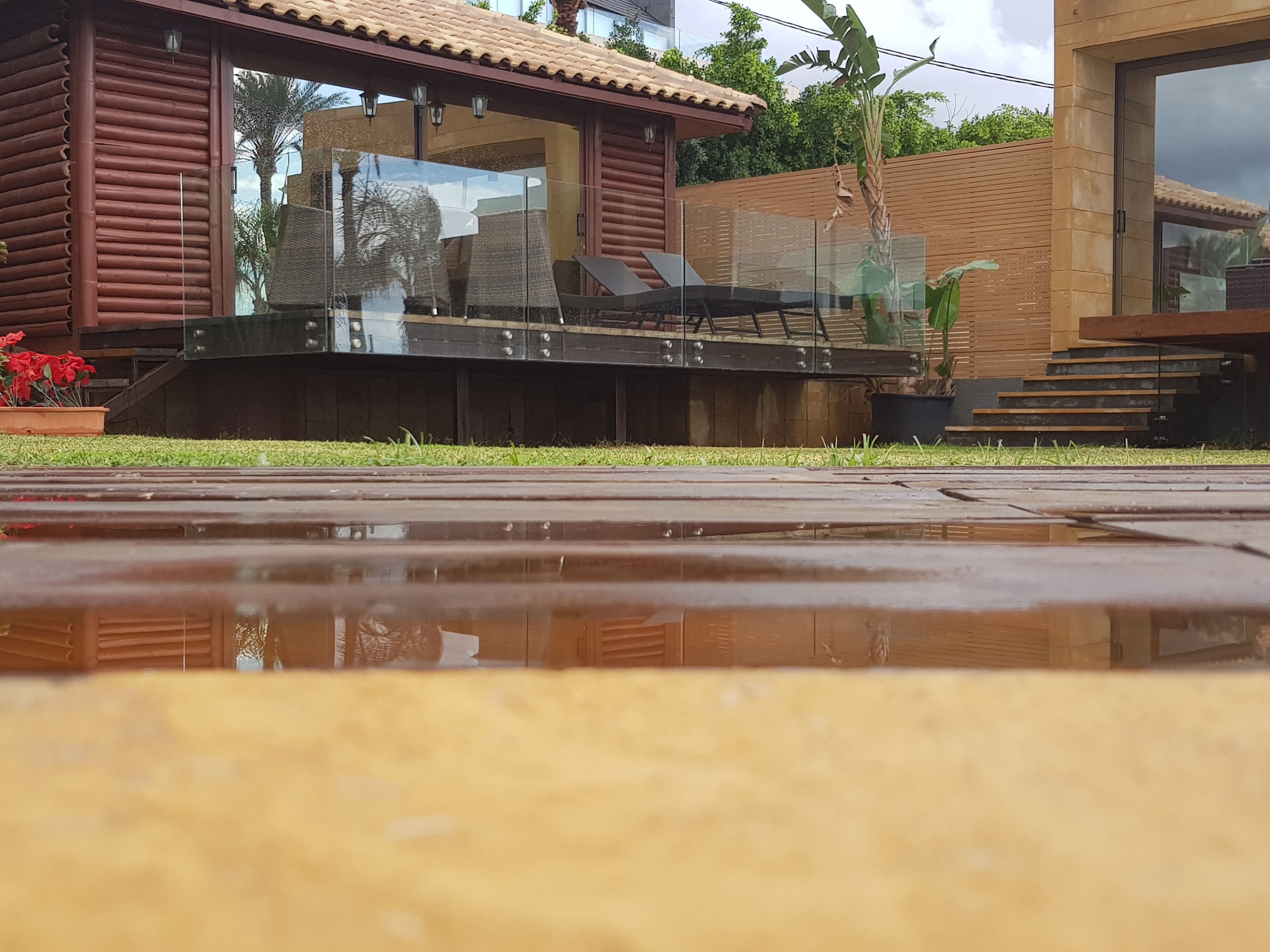 A modern house with a glass balcony, a wooden deck, and a small garden with red flowers.