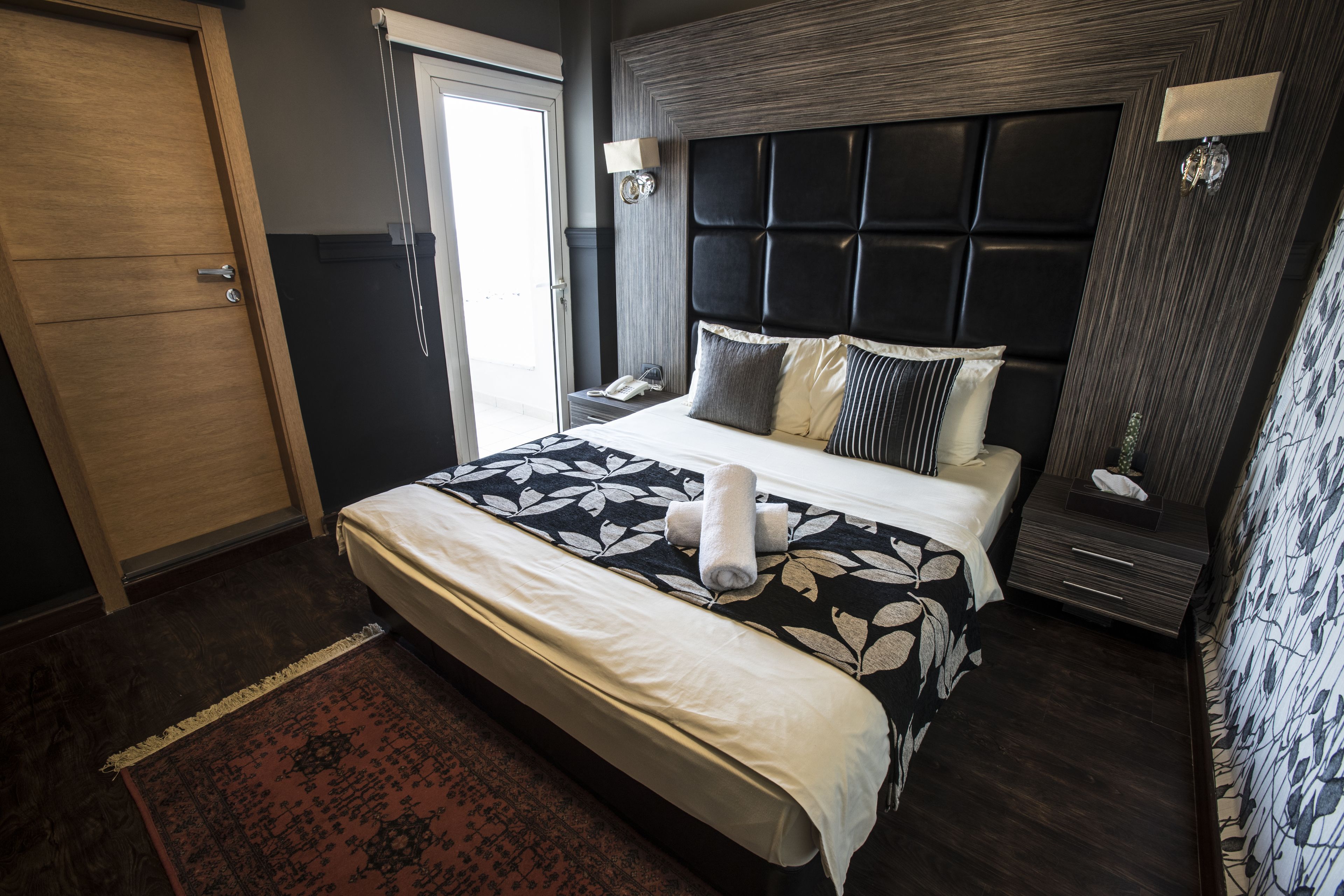 A hotel room with a large bed, a bedside table with a lamp, a wooden wardrobe, and a patterned rug.