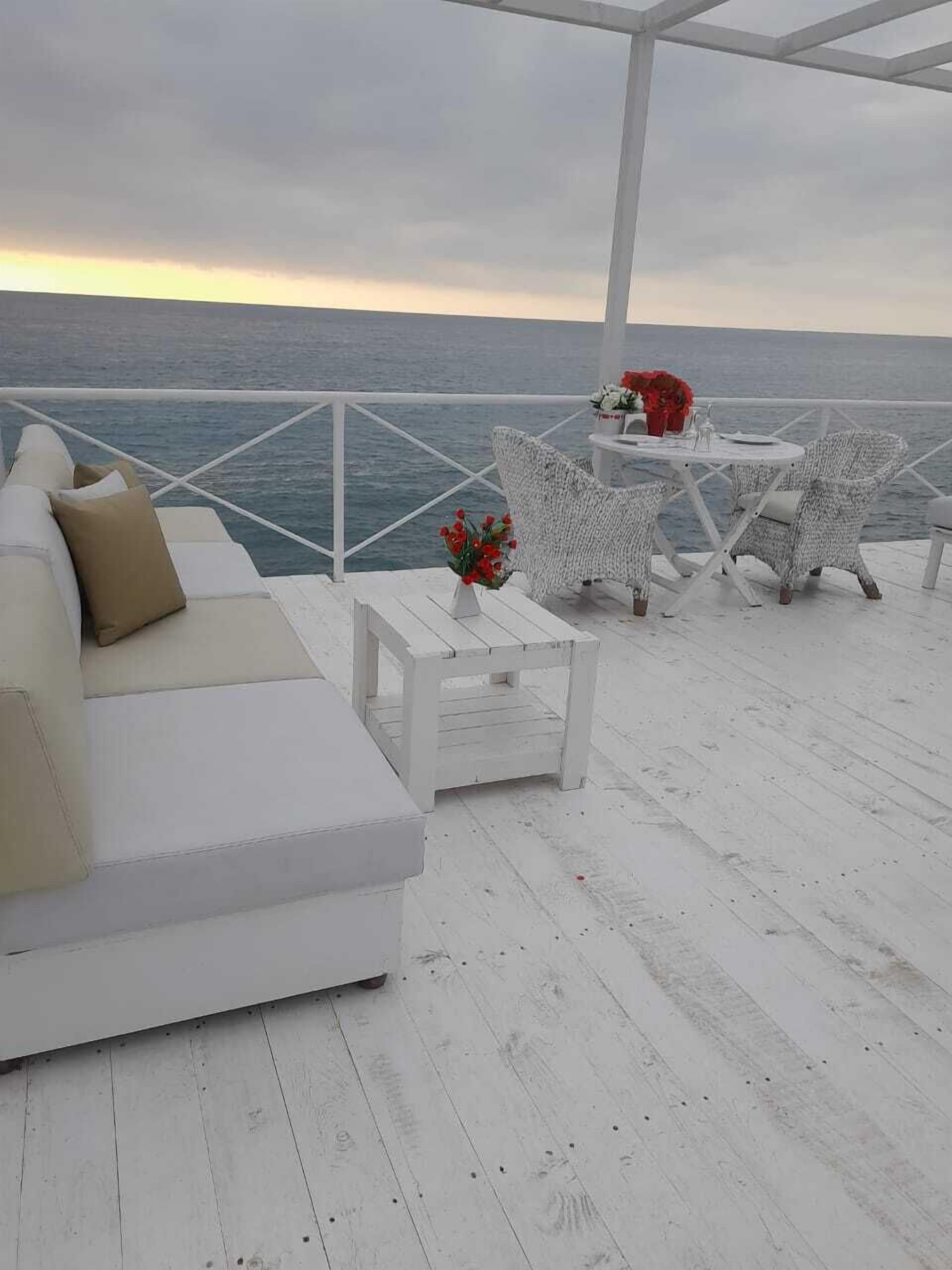 A white outdoor seating area with a view of the sea, featuring a sofa, two wicker chairs, and a small table with a vase of flowers.