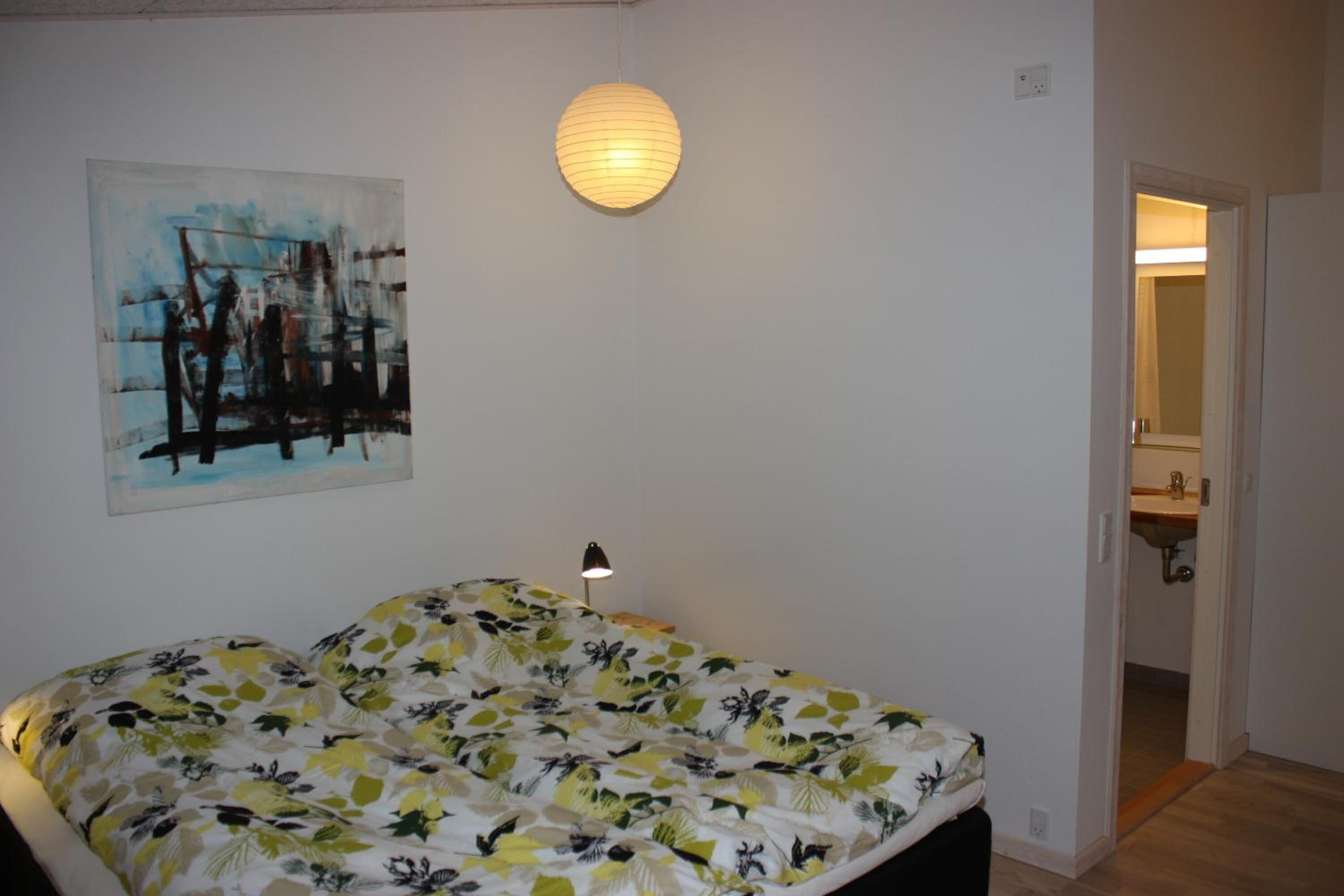 A bedroom with a bed, a wall-mounted lamp, a painting, and a door leading to a bathroom.