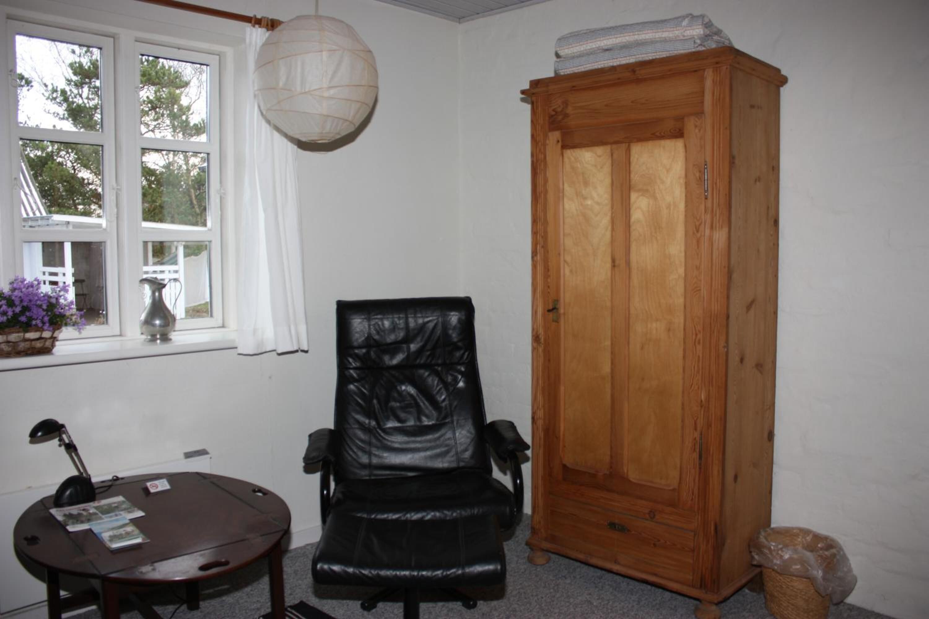 A room with a wooden cabinet, a black leather chair, a small table with a lamp, and a window with a view of trees.