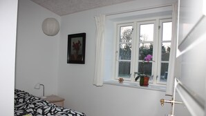 Een slaapkamer met een bed, nachtkastje, lamp, raam met gordijnen en een lijstje aan de muur.