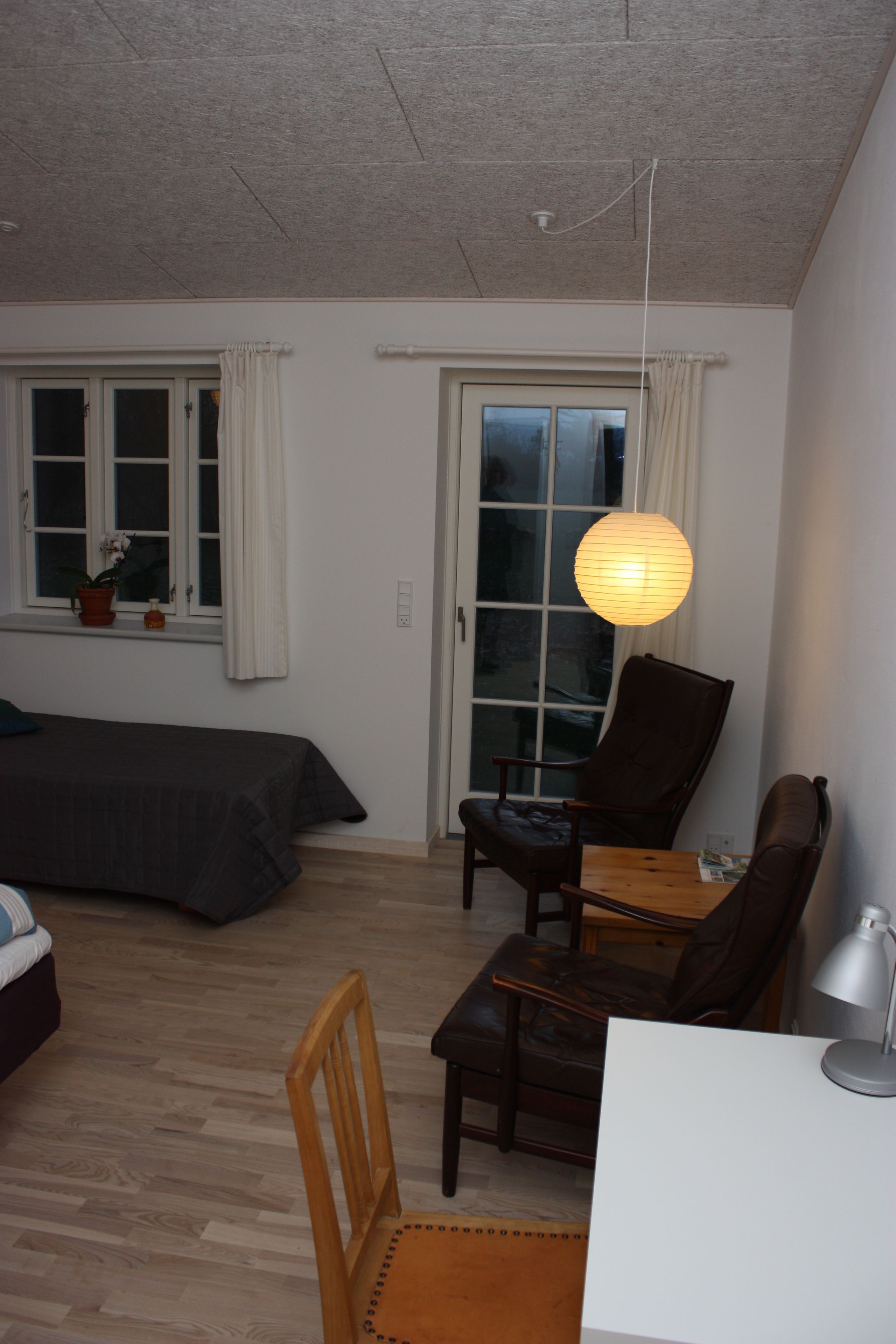 Una habitación con cama, escritorio, silla y mesa. Hay una ventana con cortinas, una planta en maceta y una lámpara colgante.