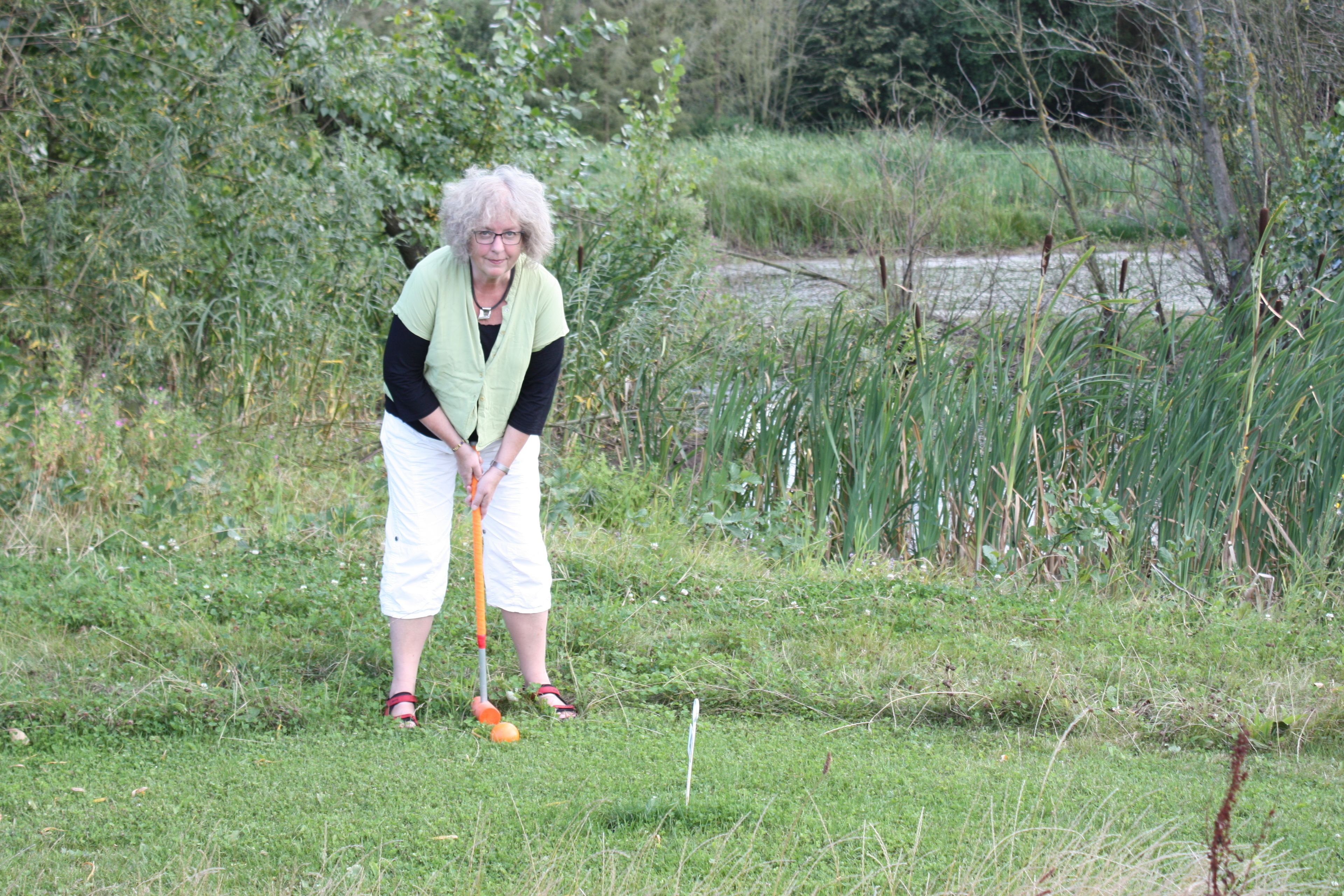 Una persona jugando croquet en un área verde al aire libre, con vegetación densa al fondo.