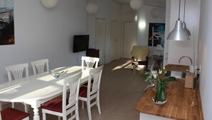 Een eetgedeelte met een witte tafel en stoelen, een keuken met een spoelbak, en een zithoek met een bank.