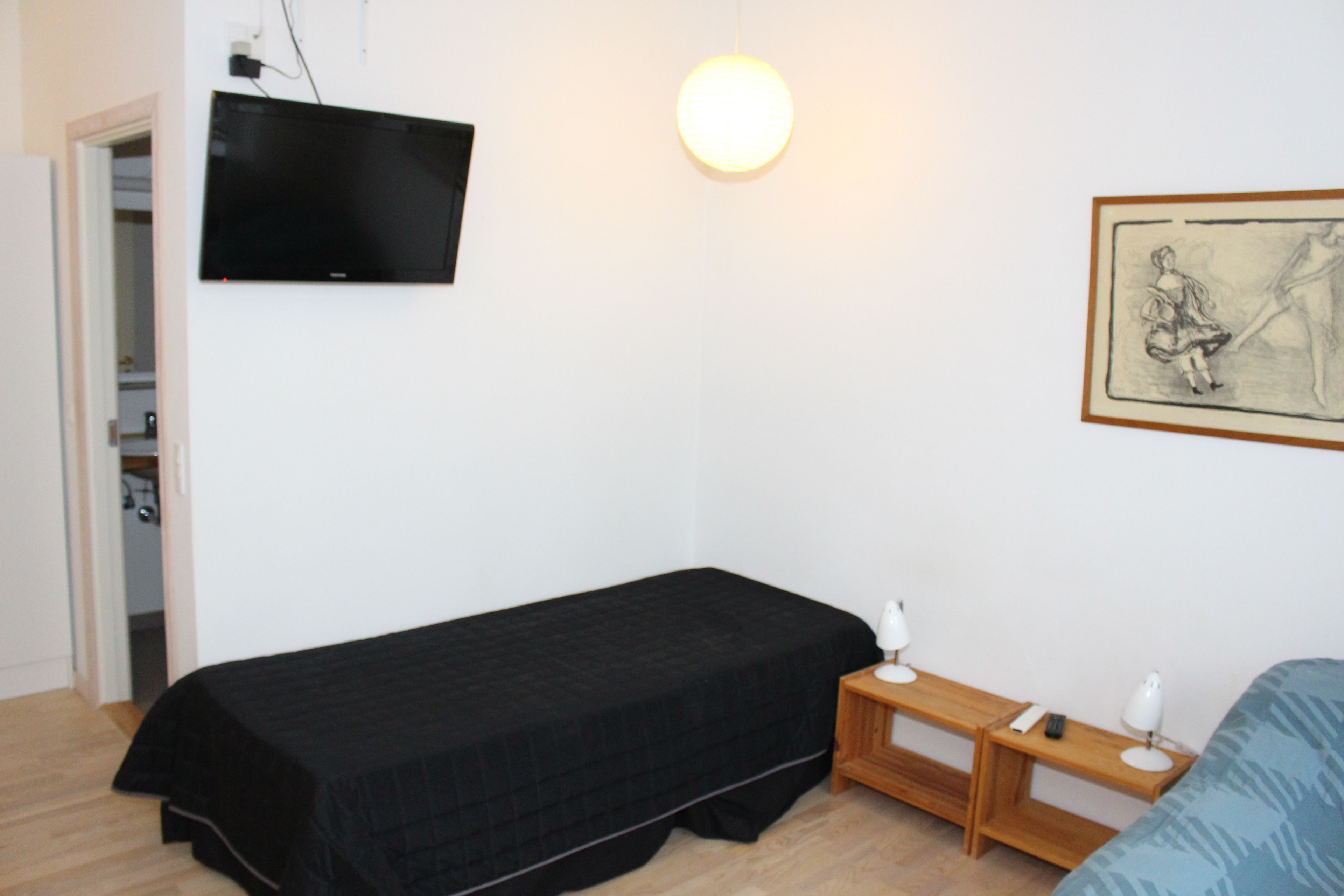 A small, single-bed room with a black bedspread, a television mounted on the wall, a bedside table with a lamp, and a framed artwork on the wall.