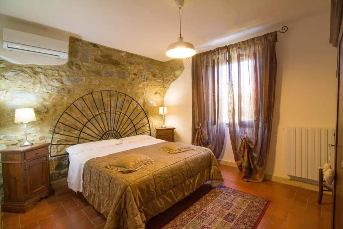 Une chambre avec un mur en pierre, un lit avec une tête de lit, une table de chevet en bois, une lampe, un radiateur et une fenêtre avec des rideaux.