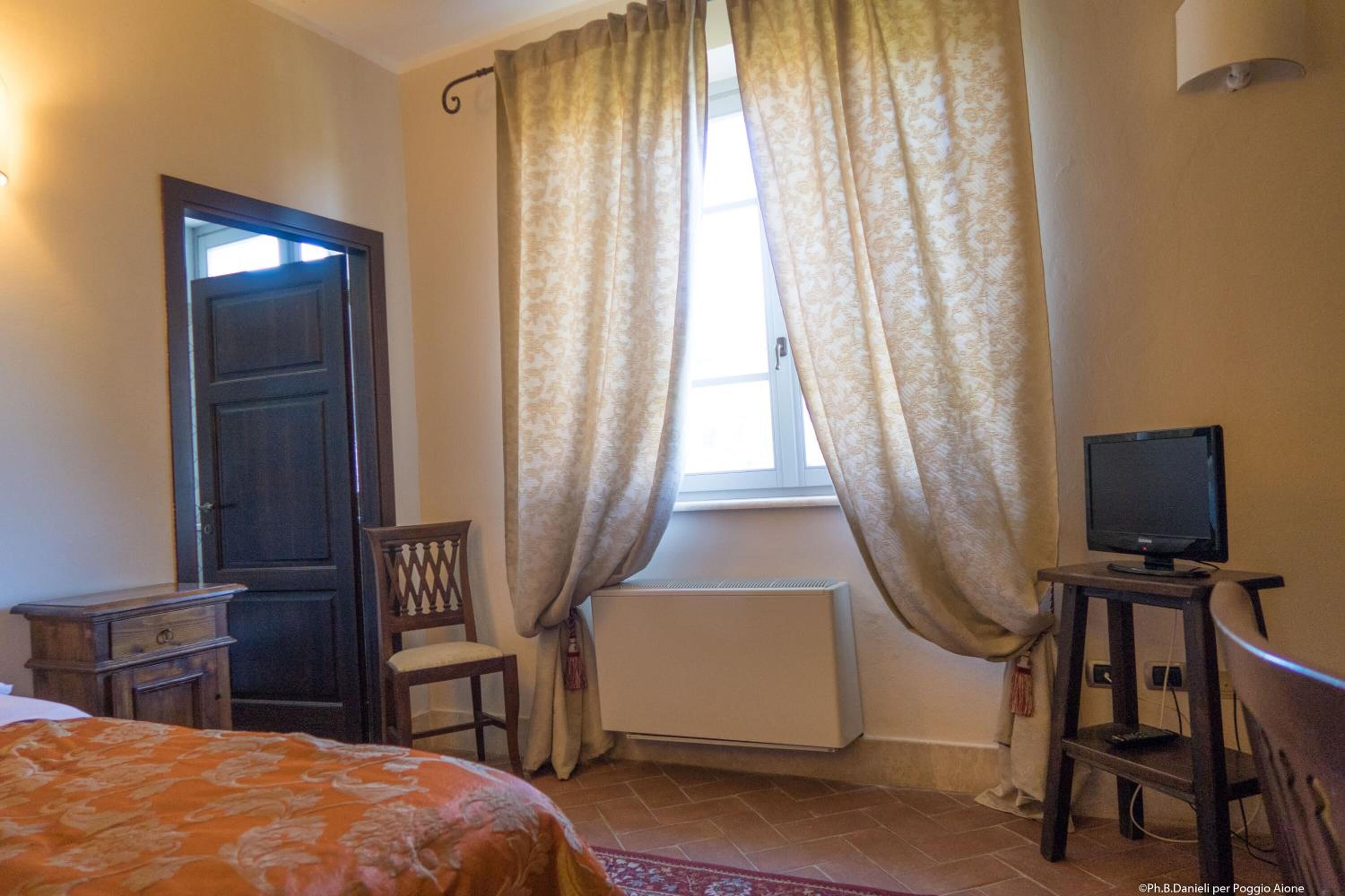Ein Schlafzimmer mit einem Bett, einem Sessel, einem kleinen Tisch mit Fernseher, einem Fenster mit Vorhängen und einem Heizkörper.