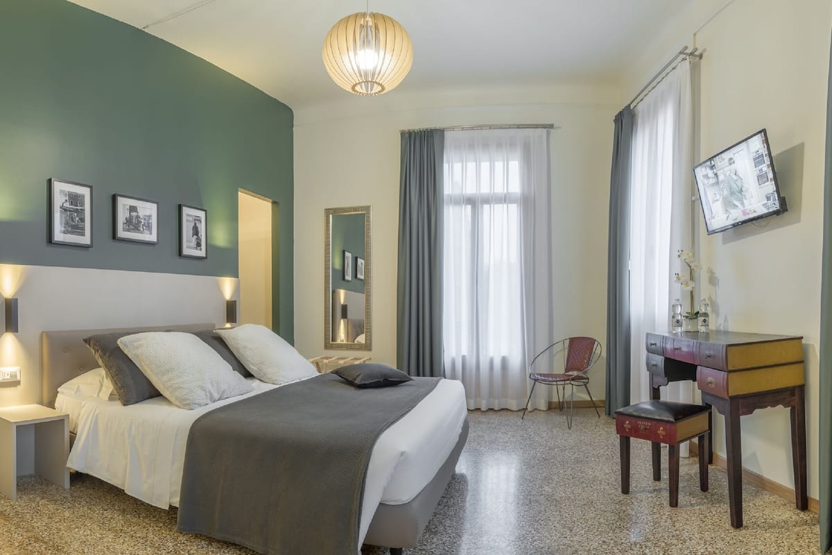 Ein Hotelzimmer mit Bett, Nachttischen, Schreibtisch, Stuhl, Spiegel, Fernseher und Vorhängen.