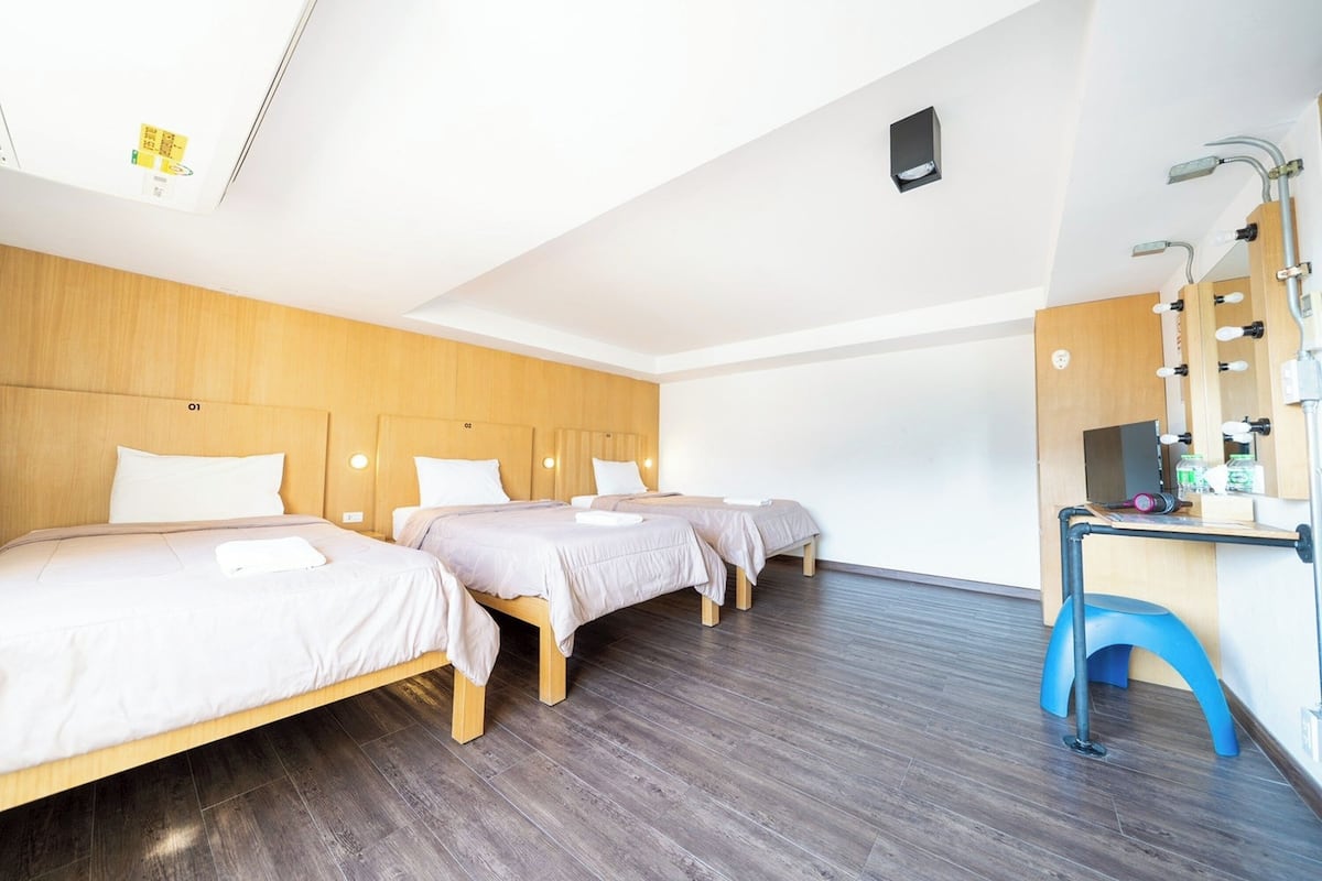 A hotel room with three beds, a wooden headboard, a desk with a mirror, and a blue stool.