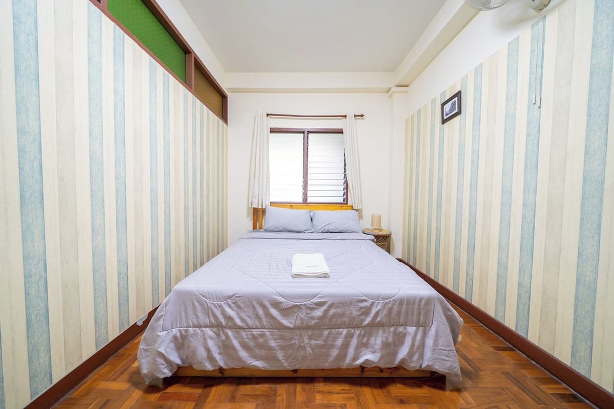 A narrow room with a single bed, striped wallpaper, and a window with curtains.