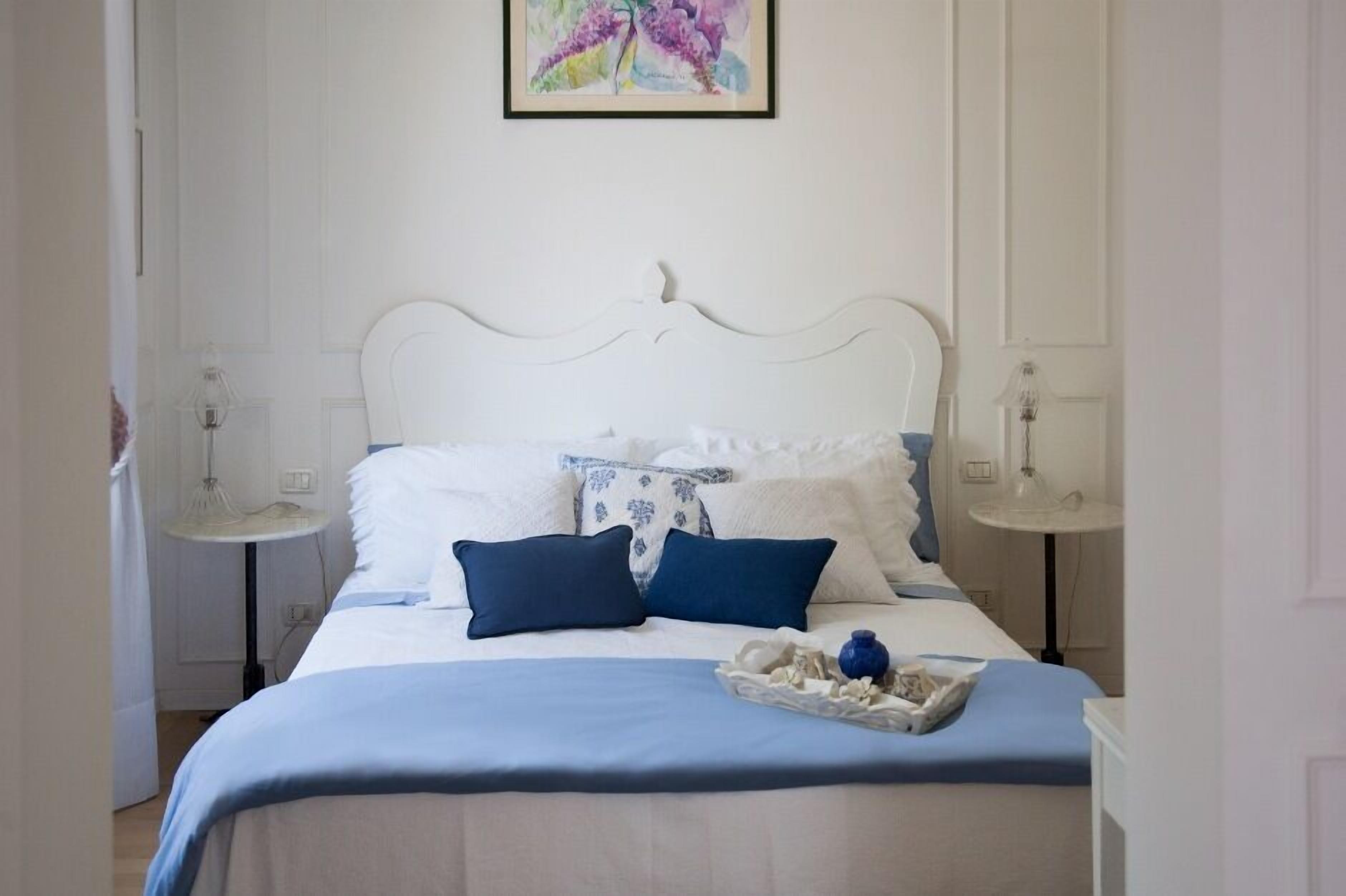 Ein ordentlich bezogenes Bett mit weißer und blauer Bettwäsche, zwei Nachttische mit Lampen sowie ein gerahmtes Kunstwerk an der Wand.