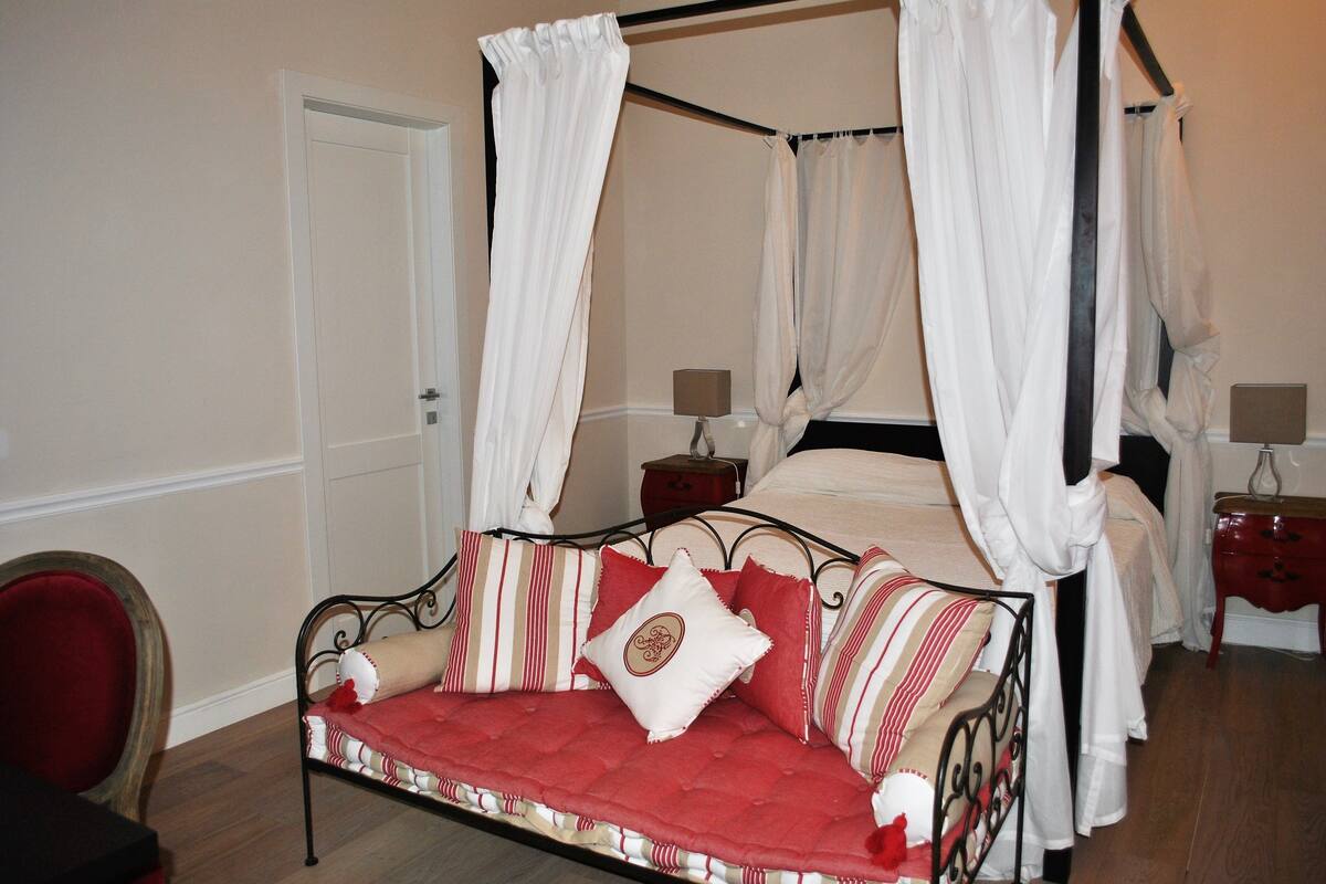 Ein Himmelsbett mit einem roten und weißen Streifenüberwurf, ein rotes, gepolstertes Sofa mit passendem Kissen sowie zwei Nachttische mit Lampen.