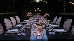 Una larga mesa de comedor dispuesta para un evento formal, con sillas a ambos lados, velas y centros de mesa con flores.
