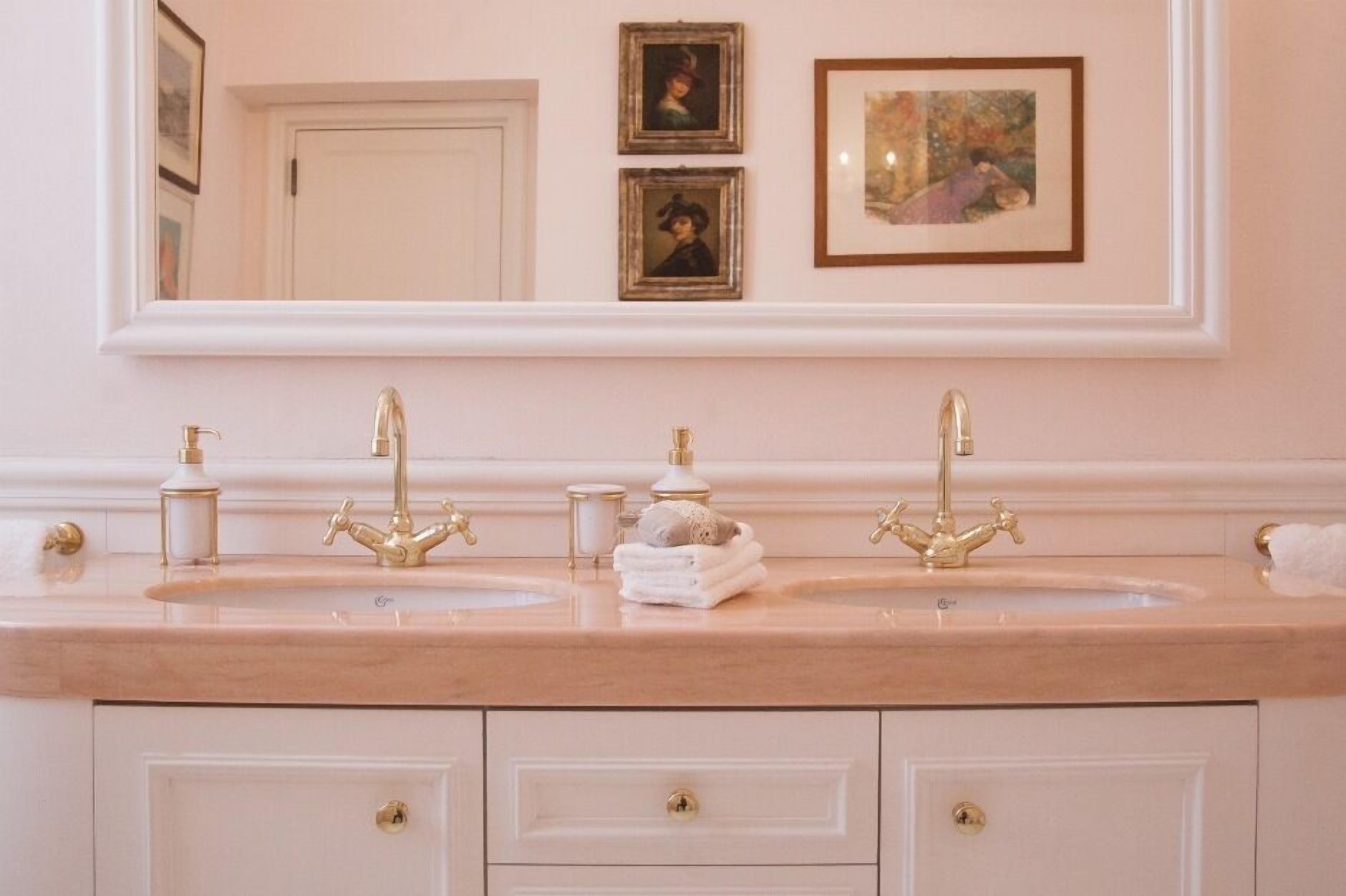A bathroom with two sinks, gold faucets, and a mirror above. There are framed pictures on the wall and a towel rack with towels.