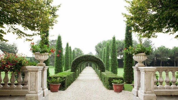 Un sendero formal de jardín con un arco central, flanqueado por setos simétricos y plantas en macetas.