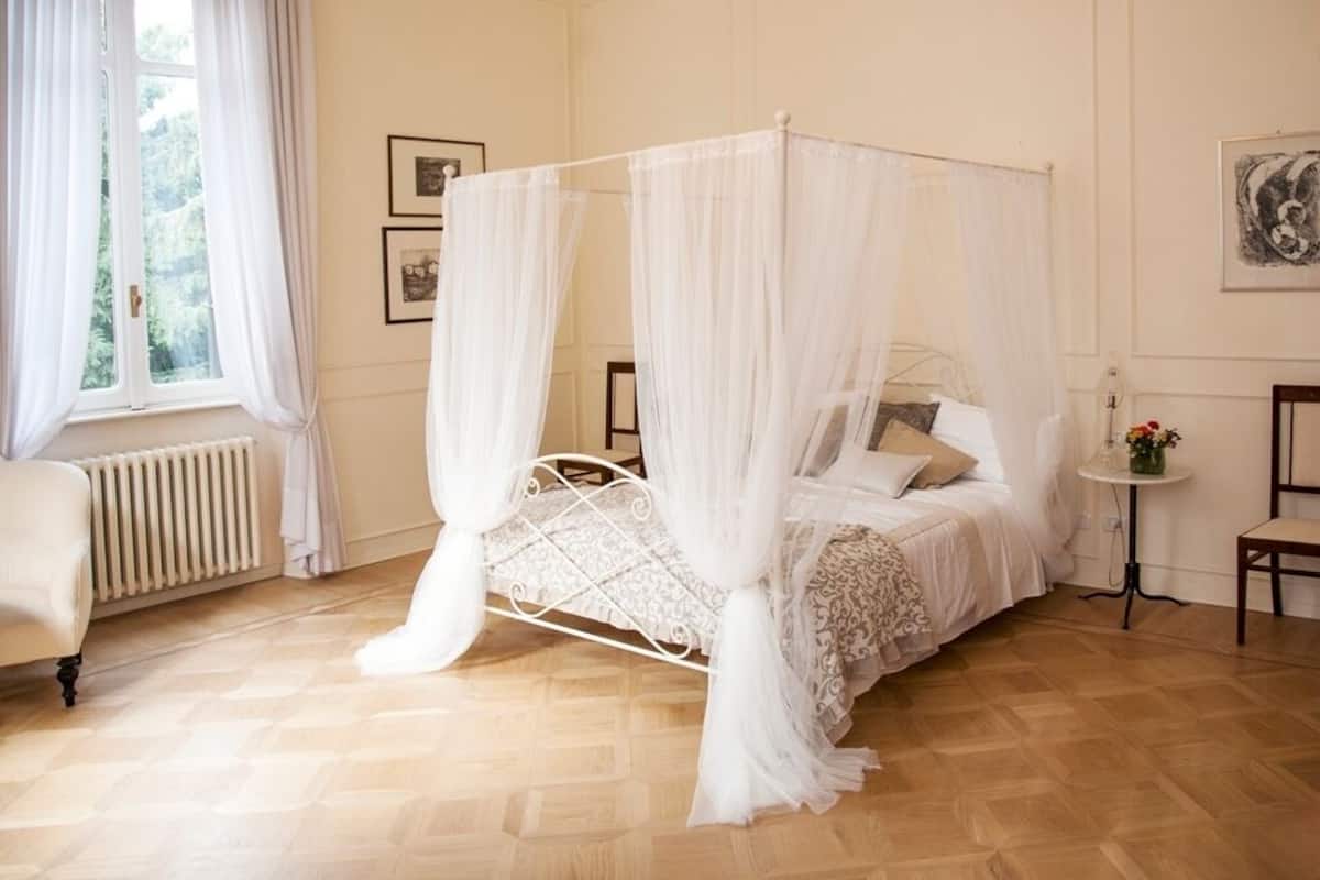 Ein Himmelsbett mit weißer Bettdecke und Kissen, eine Heizung, ein kleiner Tisch mit Blumen in einer Vase und gerahmte Bilder an der Wand.