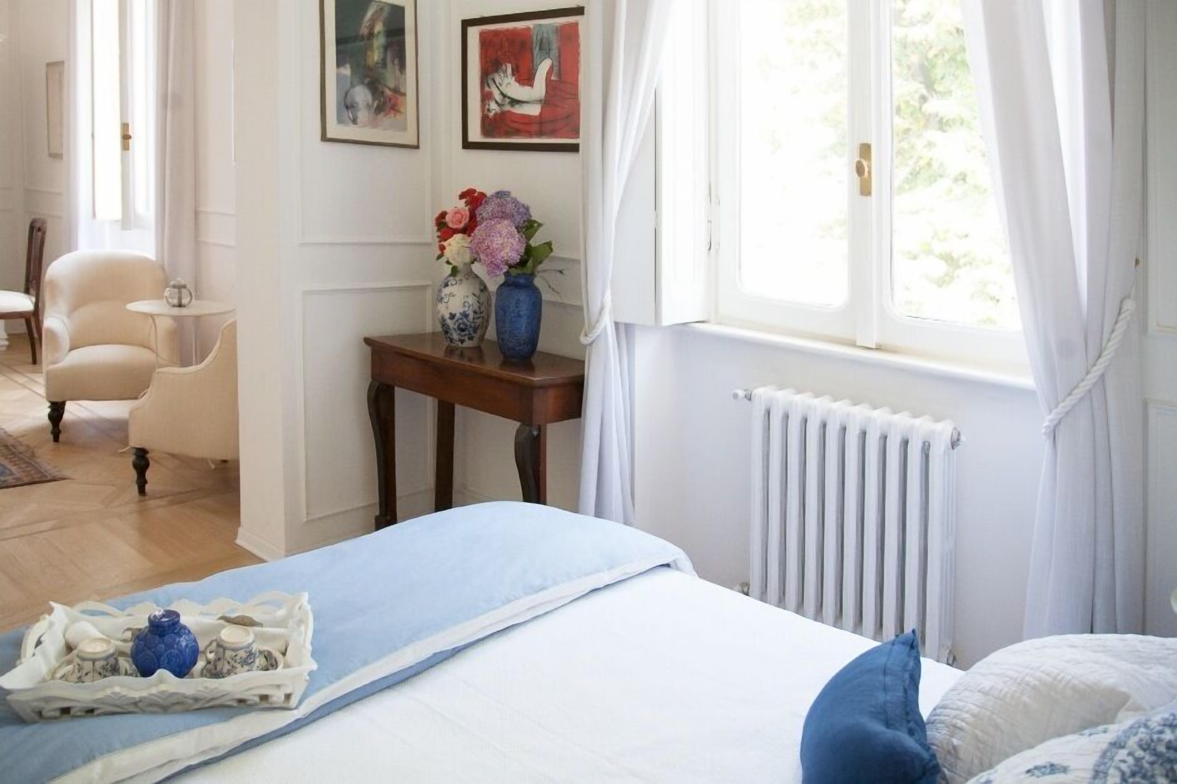 Un dormitorio con una cama, una mesita de noche con un jarrón de flores, una silla y una ventana con cortinas.