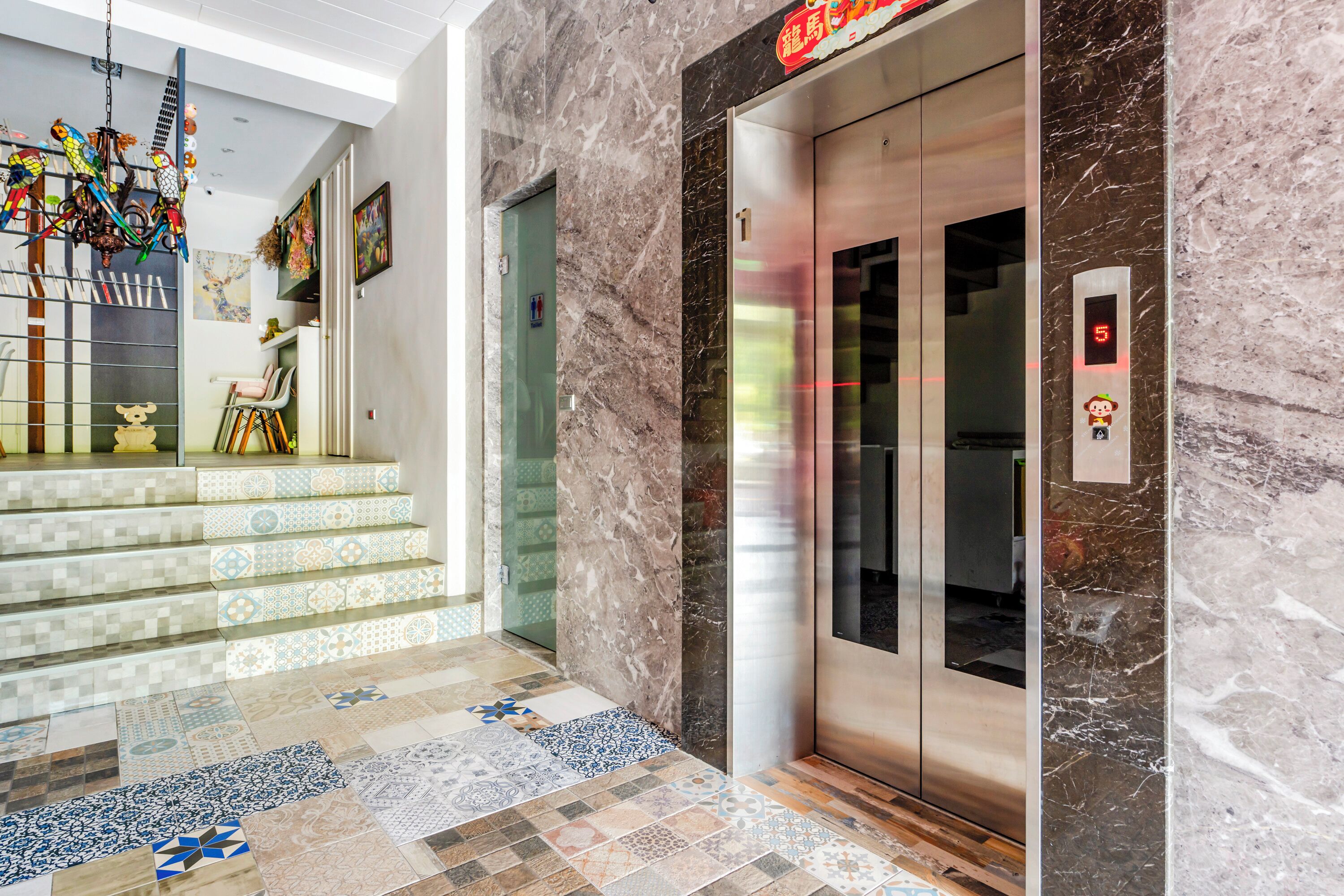 A modern lobby with a marble floor, a staircase with patterned tiles, and a glass elevator.