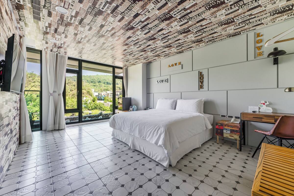 A modern hotel room with a large bed, a desk, and a view of the outdoors.