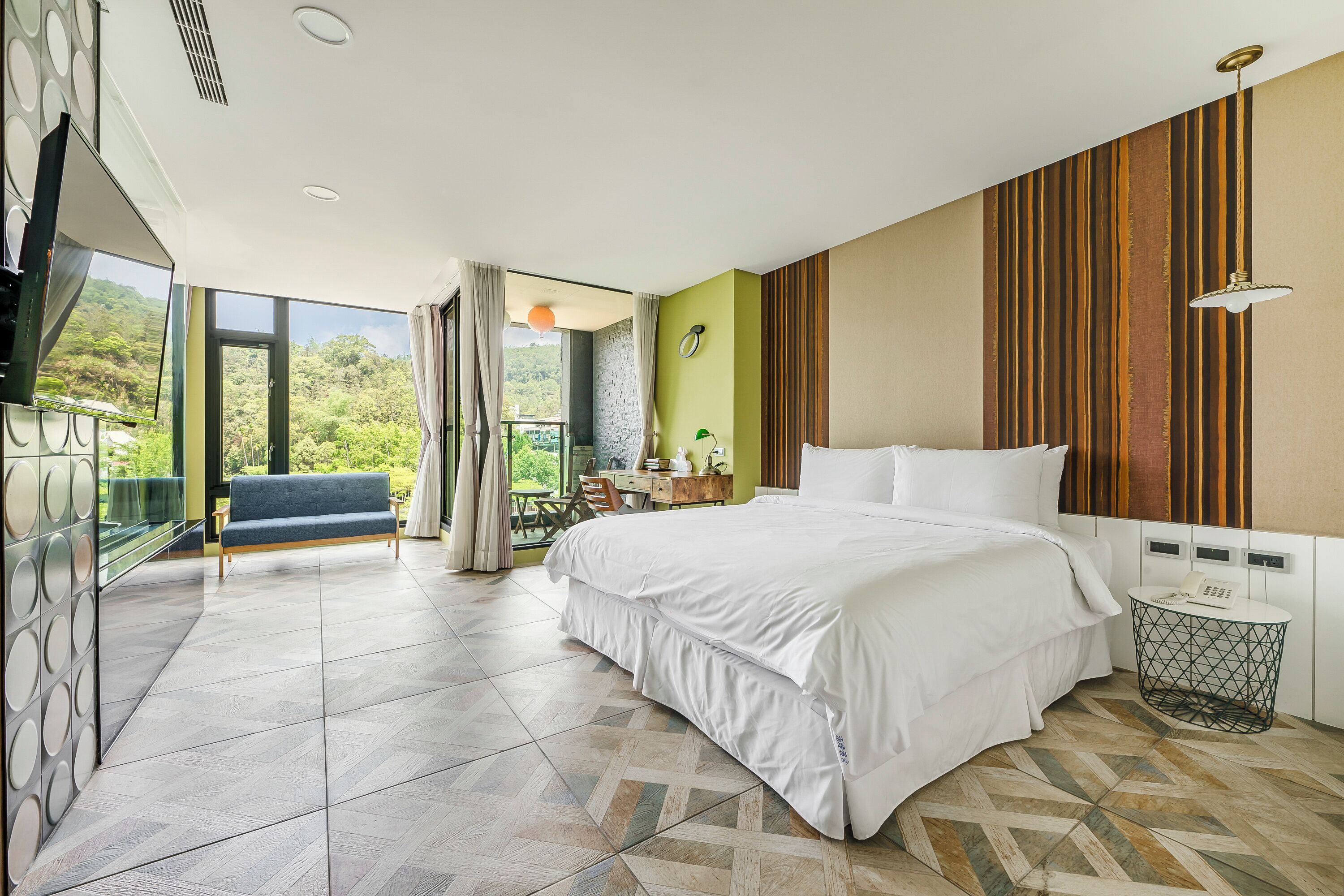 A modern hotel room with a large bed, a desk, and a balcony with a view of greenery.