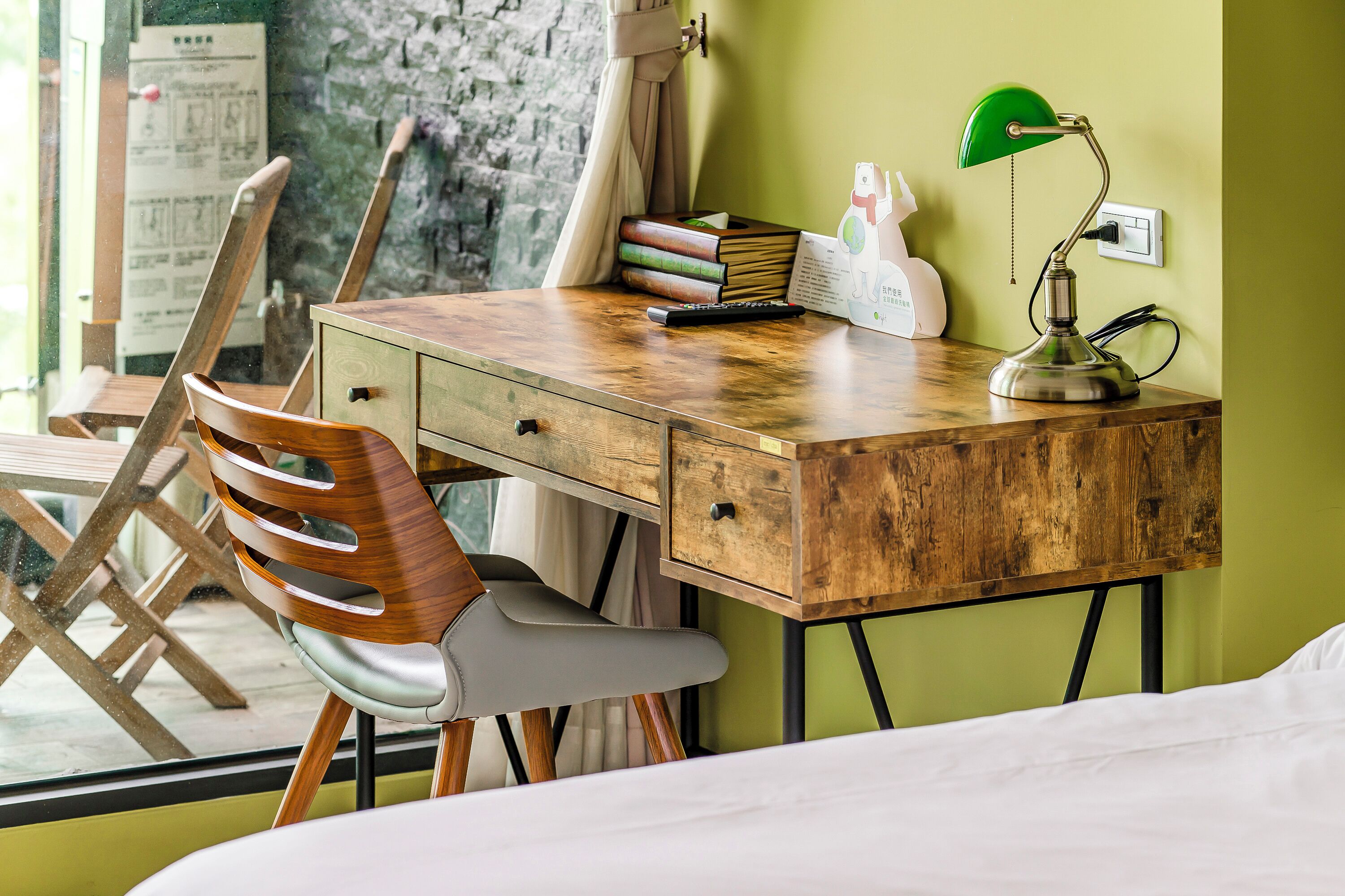 A wooden desk with a lamp, books, and a remote control. There is a chair and a bed in the room.