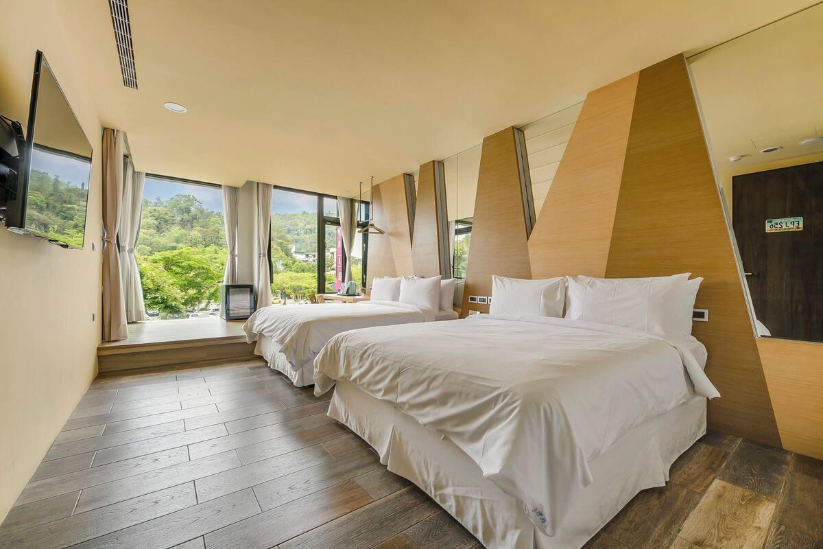 A hotel room with two beds, a TV, and large windows with a view of greenery.