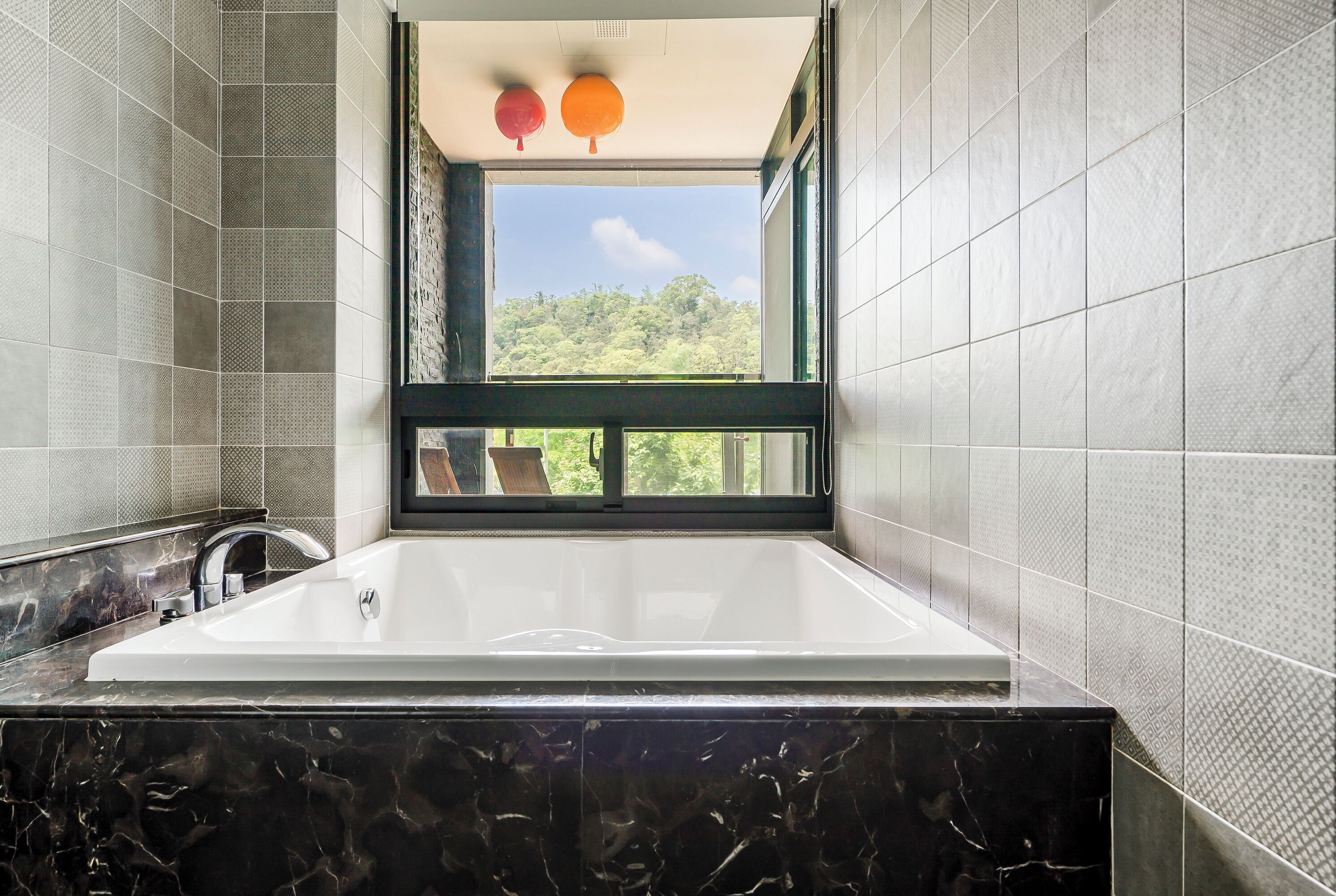 A modern bathroom with a large bathtub, a window with a view of greenery, and a black marble countertop.