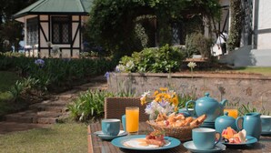 Ontbijt met eieren, spek, toast en fruit op een tafel, met uitzicht op een tuin en een gebouw op de achtergrond.