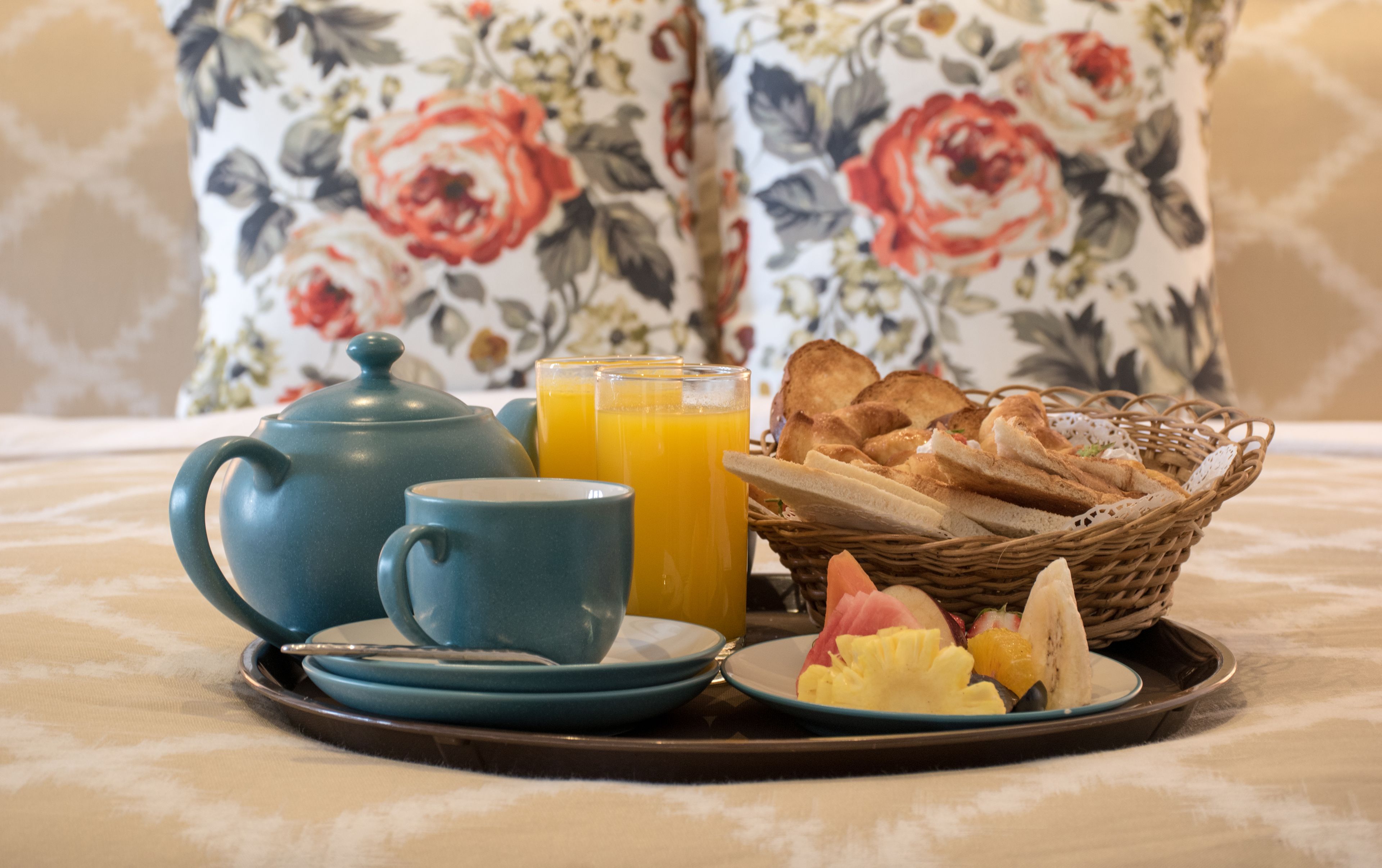 A breakfast tray with a teapot, cups, orange juice, toast, and fruit on a table with a floral-patterned tablecloth.