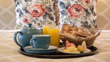 A breakfast tray with a teapot, cups, orange juice, toast, and fruit on a table with a floral-patterned tablecloth.