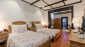 Een hotelkamer met twee bedden, houten hoofdeinden, een bureau met lamp, een telefoon en uitzicht op de gang.