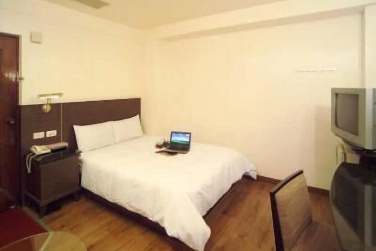 Une chambre d’hôtel avec un lit simple, une table de chevet avec une lampe, une télévision et un ordinateur portable posé sur le lit.