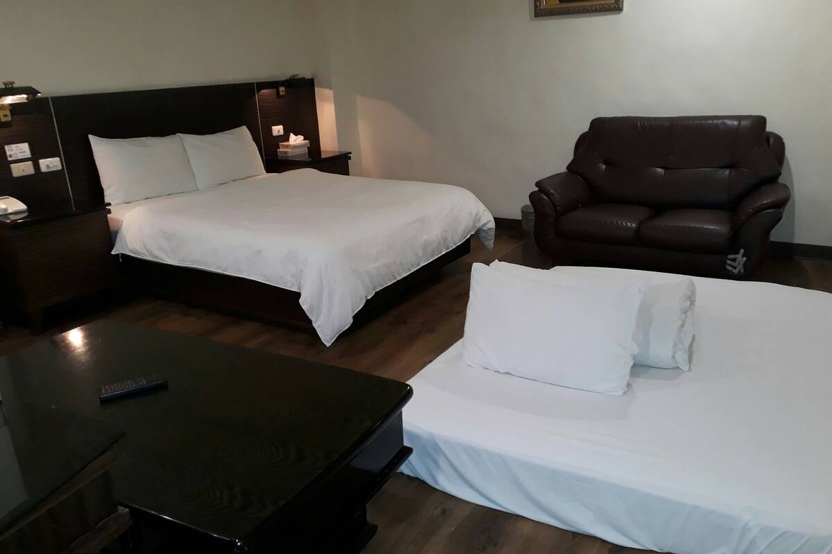 Une chambre d’hôtel avec deux lits, un fauteuil en cuir marron, une table en bois et un tableau accroché au mur.