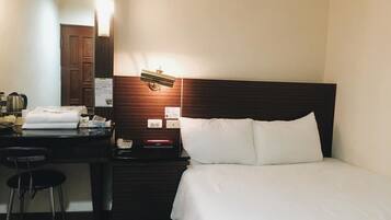A hotel room with a bed, a desk with a chair, a nightstand, a mirror, and a wall-mounted lamp.