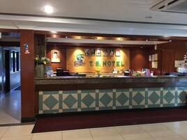 Bancada de recepção de hotel com balcão de check-in, quadros nas paredes e uma placa que diz 'F.T.S. HOTEL'.