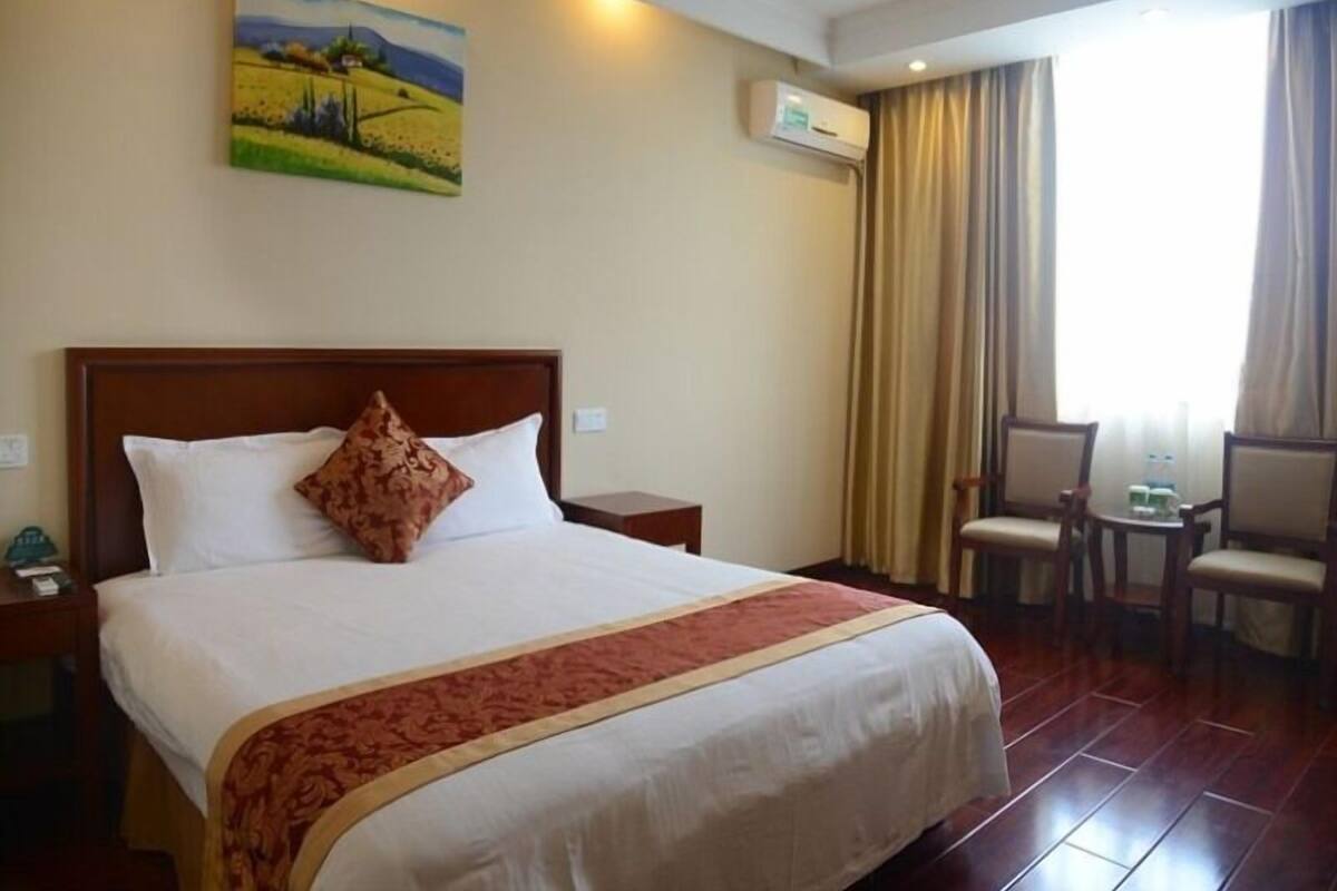 Een hotelkamer met een groot bed, een schilderij aan de muur, een airconditioningapparaat, een raam met gordijnen en een kleine tafel met stoelen.