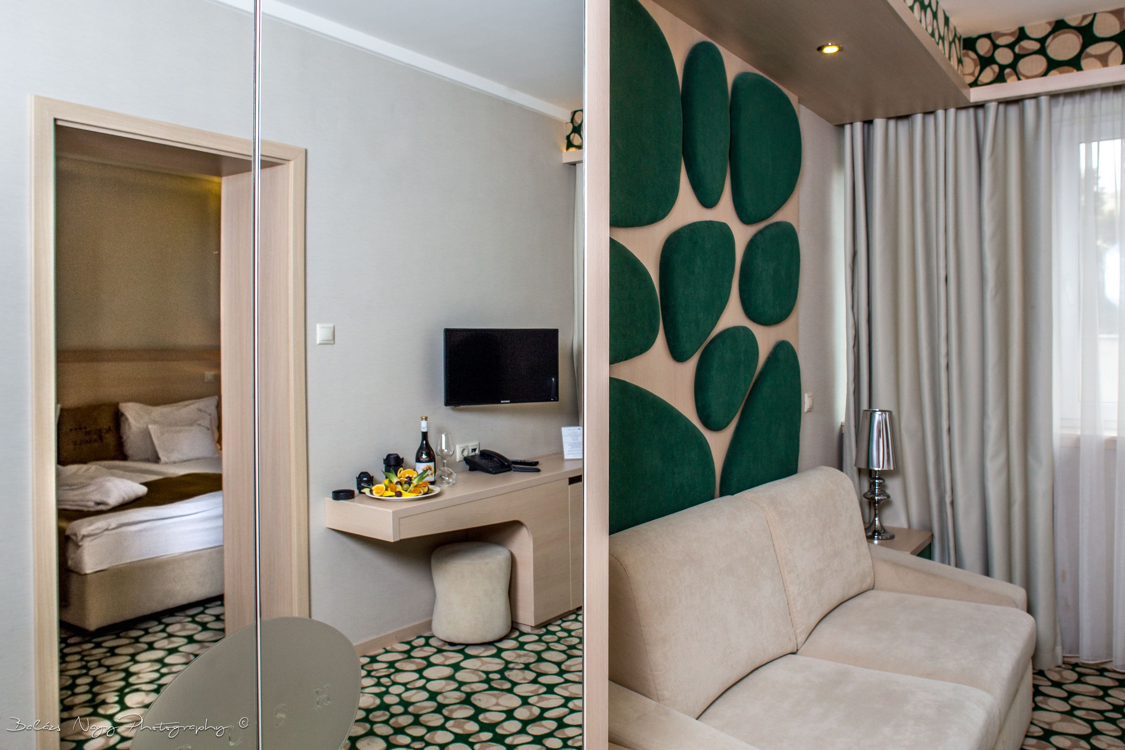 A hotel room with a bed, a desk with a TV, a sofa, and a patterned floor.