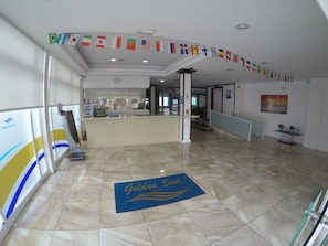 Un hall d’hôtel comprenant un comptoir d’accueil, un paillasson et un mur décoré de drapeaux internationaux.