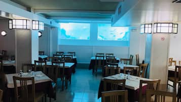 Restaurante com várias mesas e cadeiras, uma janela grande com vista e luminárias na parede.