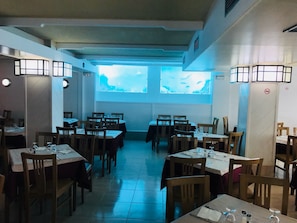 Un restaurant doté de nombreuses tables et chaises, d’une grande fenêtre offrant une belle vue et de luminaires fixés aux murs.