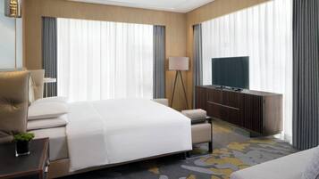 A hotel room with a large bed, a TV, a desk with a lamp, and a view of the city through large windows.