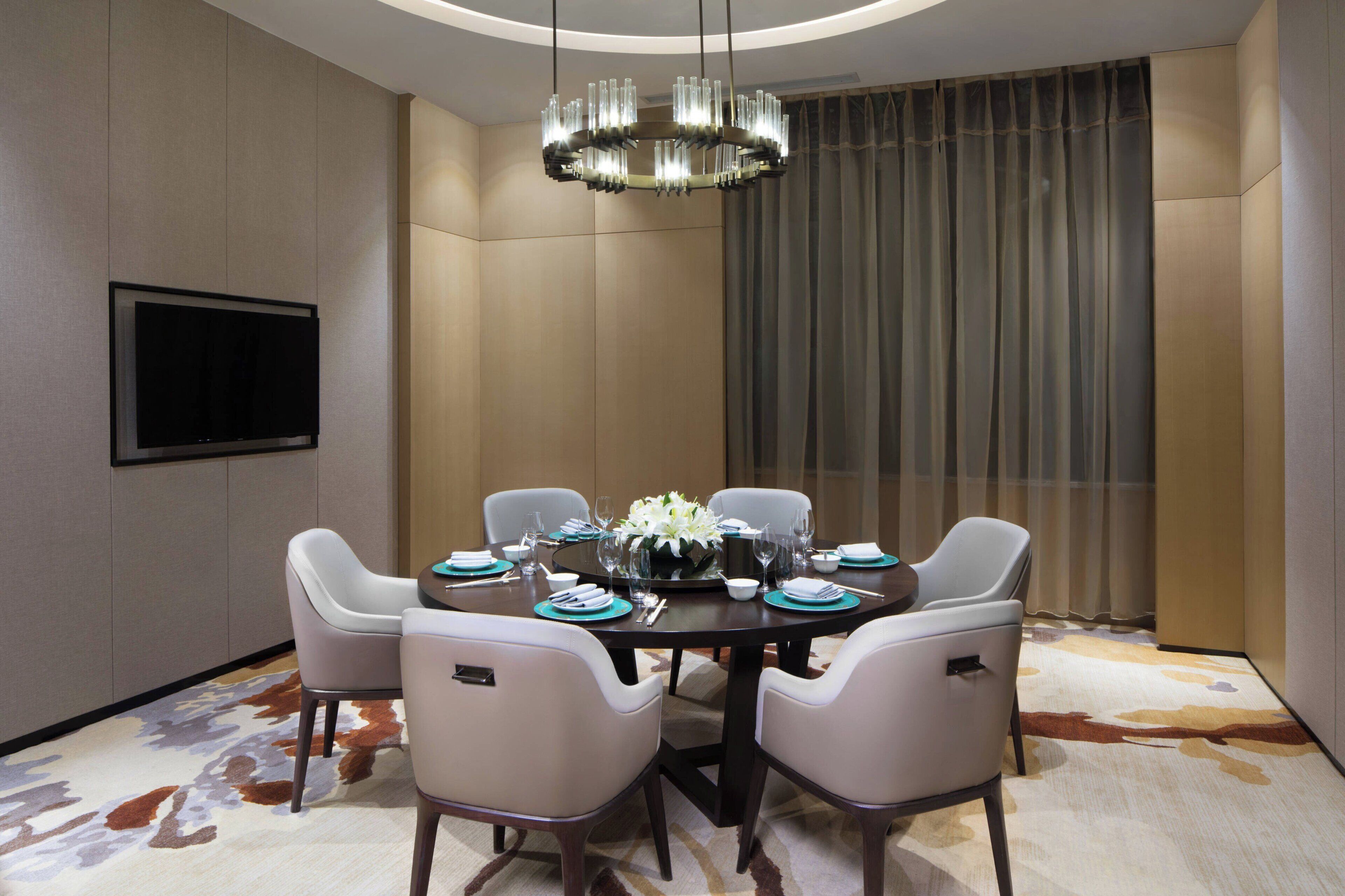 A modern dining room with a round table, six chairs, a chandelier, a flat-screen TV, and a large window with curtains.