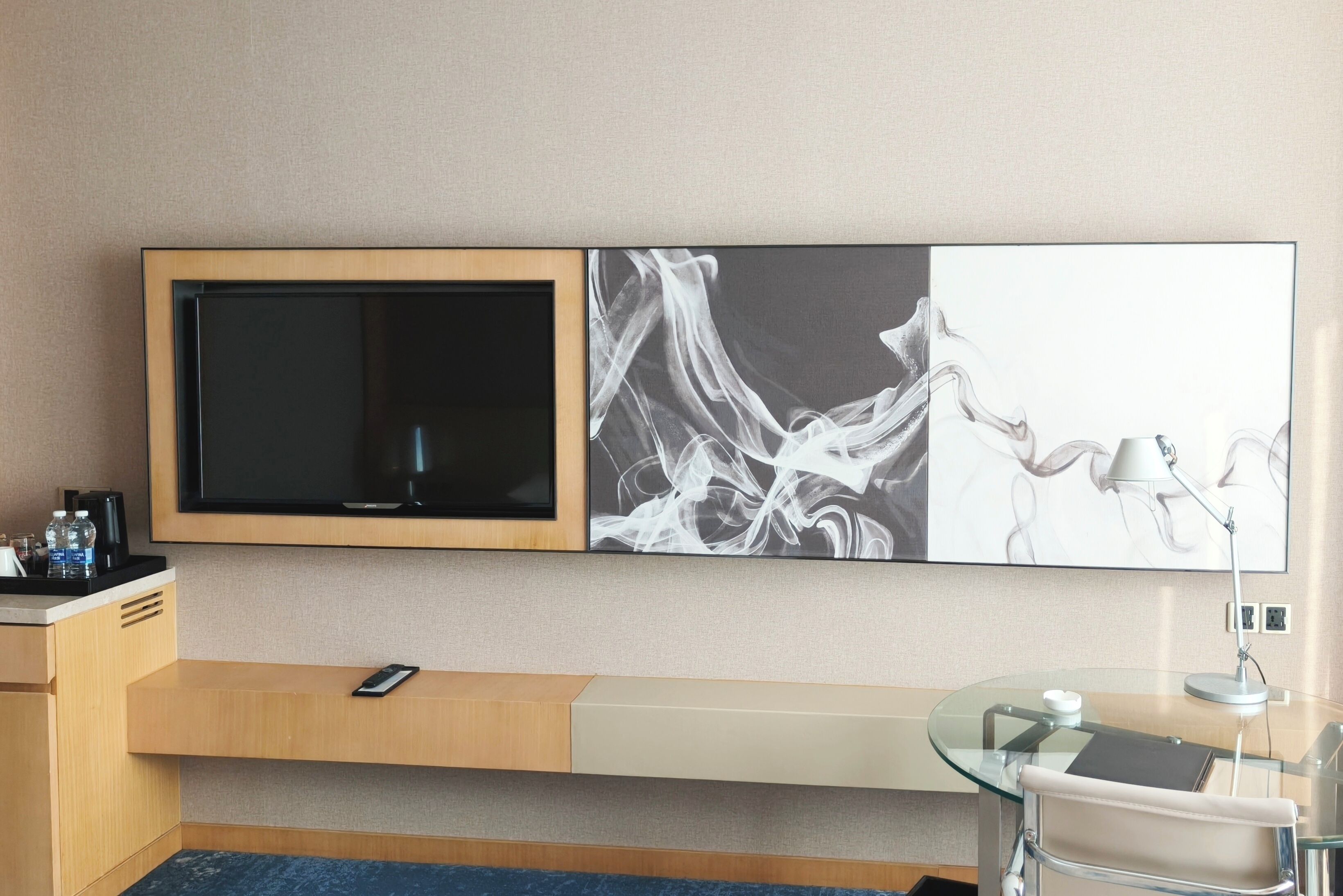 A modern hotel room with a large flat-screen TV, a glass desk with a laptop, a chair, and a wall-mounted shelf with a phone.