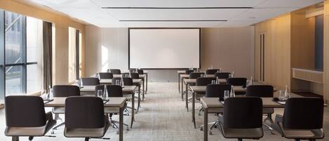 Una sala conferenze con file di tavoli e sedie, uno schermo per proiettore e ampie finestre.