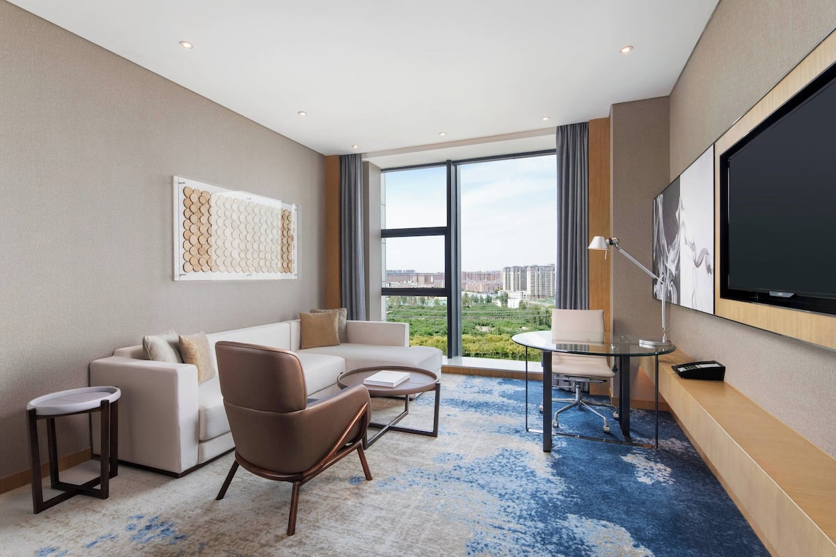 Une chambre d’hôtel moderne dotée d’une grande fenêtre, d’un canapé, d’un fauteuil, d’une petite table et d’un téléviseur fixé au mur.