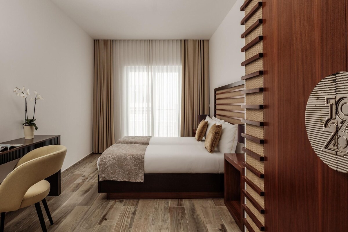 A hotel room with a bed, a desk, a chair, a potted plant, and a large window with curtains.