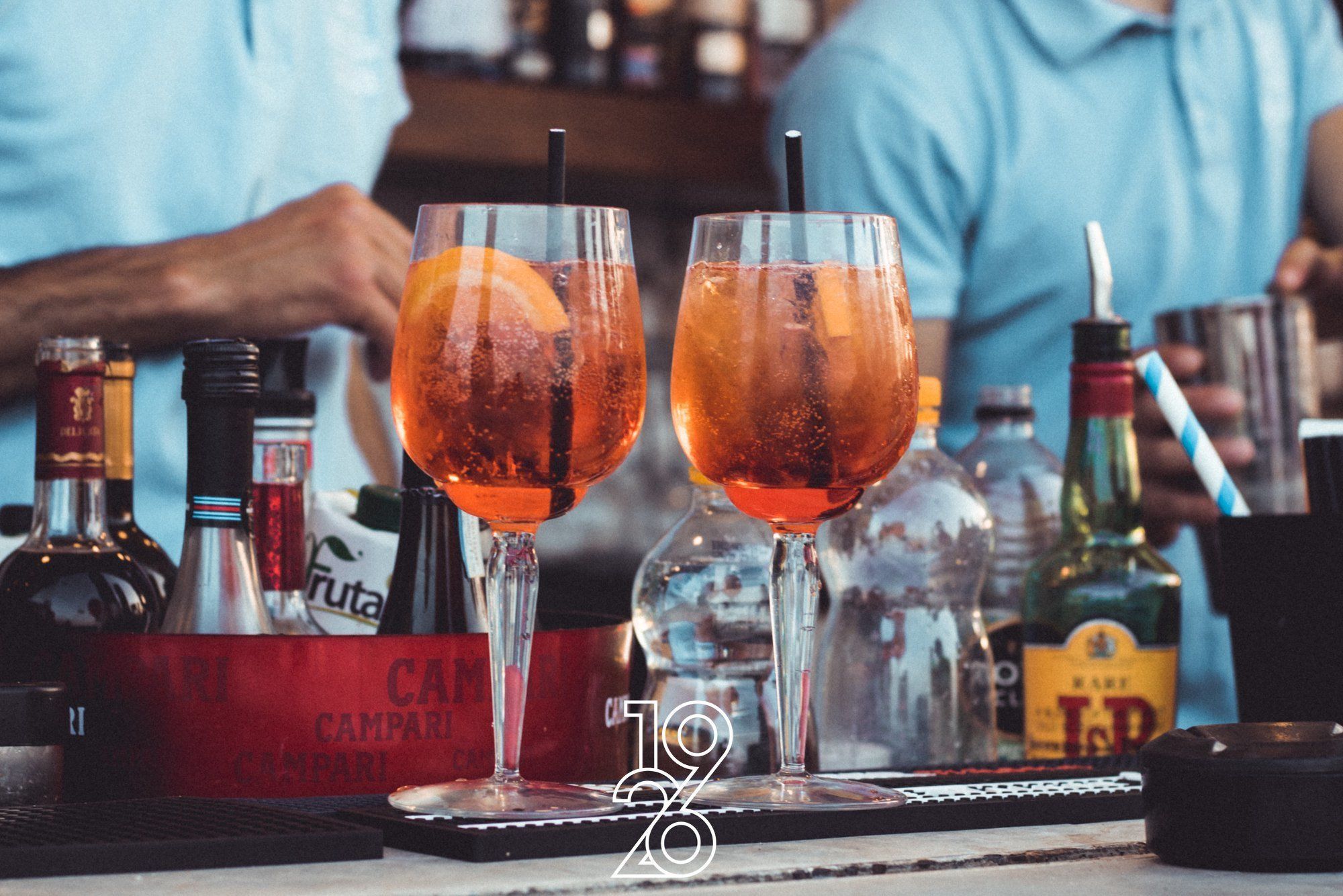 Two glasses of amber-colored cocktails on a bar counter with various bottles and a branded coaster.
