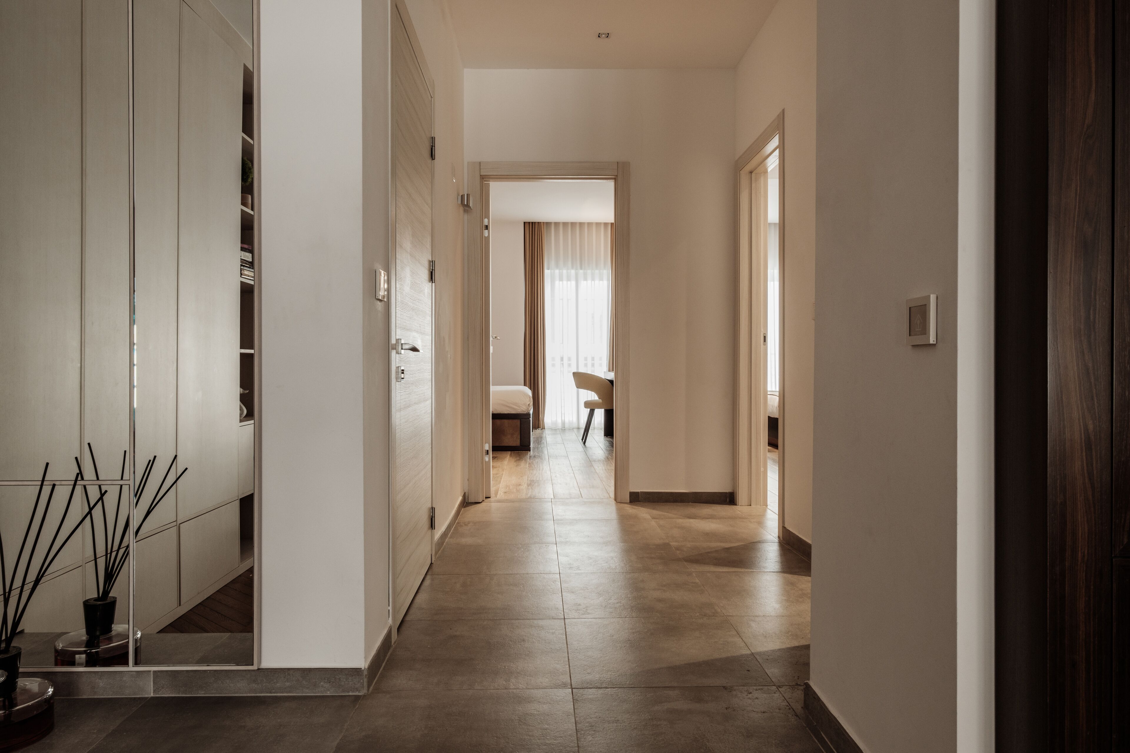 A modern hotel hallway with a neutral color palette, a large mirror, and a view into a room with a bed and curtains.