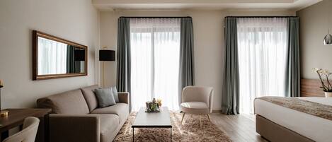 A modern hotel room with a sofa, armchair, and a bed. There is a coffee table with a decorative item, and the room has large windows with curtains.